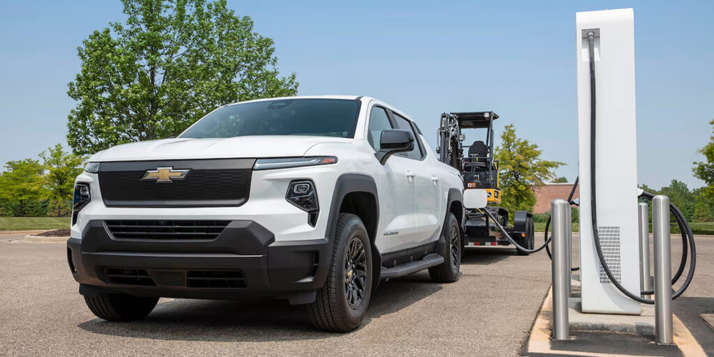 Photo of a 2026 Chevrolet Silverado EV, with trailer carrying a small construction vehicle attached, connected to a charging station.